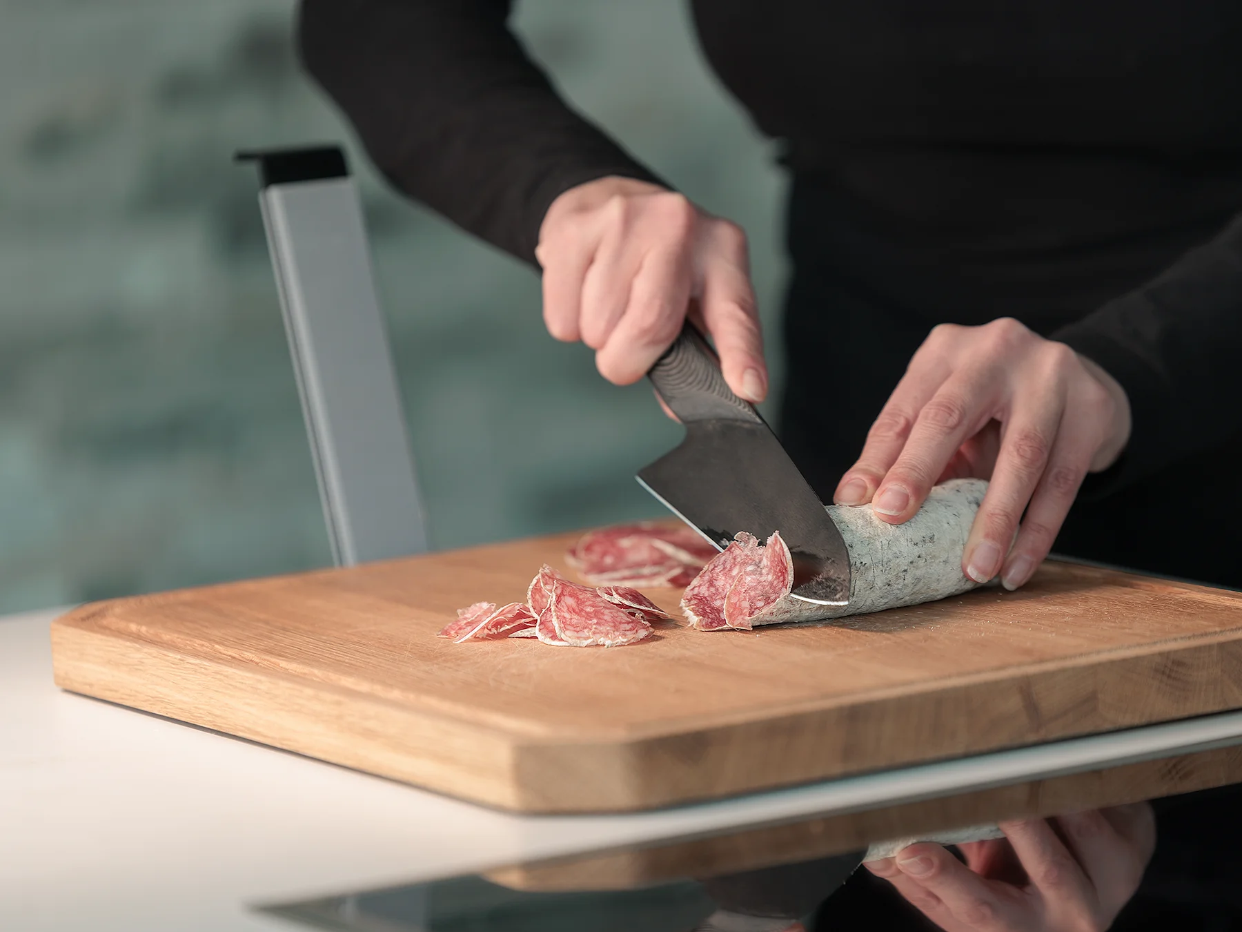 Person schneidet Salami mit einem TYROLIT LIFE Darkline Fly Wheel Cut S auf dem WOLFBOARD.life Eichenholzbrett in einer modernen Küche